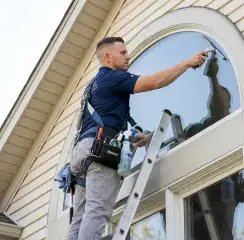 Professional window cleaner on a ladder using a squeegee to clean an exterior arched window, Wichita, KS.